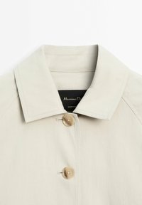 Beige jas met grote spitse kraag en twee ronde beige knopen die aan de voorkant zijn vastgemaakt.