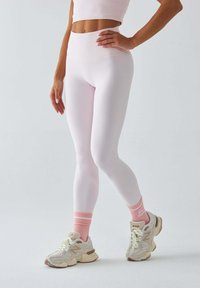 Jasnoróżowe legginsy z wysokim stanem, w zestawieniu z białymi i beżowymi sneakersami z różowymi akcentami oraz skarpetkami w paski. Gładka faktura.