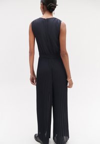 Zwarte geplooide jumpsuit met een mouwloos ontwerp, elastische tailleband en verborgen ritssluiting aan de achterkant. Vloeiende, soepele stof met een relaxte pasvorm.