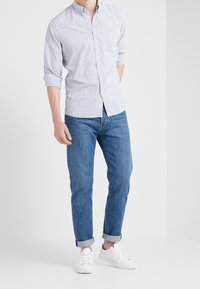 Hombre con una camisa de botones a rayas en azul claro con las mangas arremangadas, jeans azules con dobladillo y zapatillas blancas de pie contra un fondo blanco.