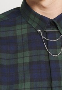 Camisa de cuadros con tonos verde oscuro y navy, un patrón texturizado, cierre de botones y una cadena decorativa de plata cruzando el collar.