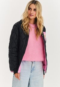 Blondýna v černé prošité bundě, otevřené přes růžový huňatý svetr a světle modré džíny s vysokým pasem, na jednobarevném pozadí.