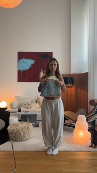 Denim kratek top, ki ga drži oseba, v zbledeli modri barvi. Sivi trenirki, bele nogavice in nevtralno obarvana dnevna soba z lampo.