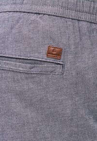 Κοντινό πλάνο τσέπης από γκρι υφασμάτινο παντελόνι με μικρή καφέ δερμάτινη ετικέτα ραμμένη πάνω από το άνοιγμα της τσέπης.