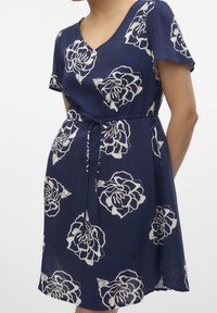 Marinblå klänning med korta ärmar, V-ringning och vit blommig mönstring. Inkluderar ett knytbälte i midjan och har ett svävande, lätt tyg.