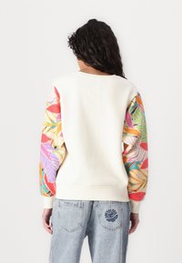 Sudadera color crema con mangas vibrantes y estampadas que presentan hojas tropicales. Diseño en azul en el bolsillo trasero de unos jeans de mezclilla clara.