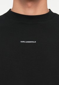 Melna kokvilnas T-krekls ar ribotu uzdinošo apkakli, kam ir balts izšūts "KARL LAGERFELD" logo uz krūtīm. Gluda tekstūra.