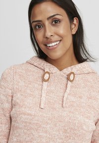 Svetlo roza pleteni kapucar z teksturiranim vzorcem, ki vključuje vrvico in okrogle lesene detajle na izrezu.