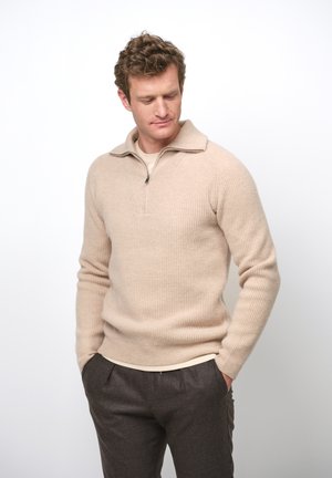 Beige geribbelde trui met een halve rits op de kraag, gedragen over een lichte top, gecombineerd met een paar bruine broeken. Kenmerkt zich door een fijne textuur en atletische pasvorm.