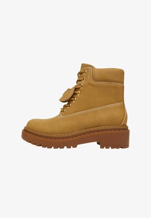 Stivaletto alla caviglia in pelle color cuoio con suola spessa a carrarmato, collo imbottito e occhielli in metallo per i lacci coordinati color cuoio, mostrato in profilo laterale.