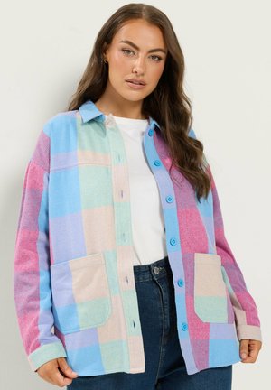 Femme aux longs cheveux bruns portant une veste à carreaux pastel avec des boutons bleus, une chemise blanche et un jean bleu foncé sur un fond uni.