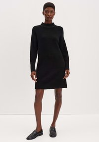 Schwarzes Strickkleid mit hohem Ausschnitt, langen Ärmeln und strukturiertem Finish. Oberschenkellang mit geradem Schnitt und dezenten Rippungen an den Ärmeln.