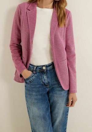 Mujer que lleva un blazer rosa ajustado sobre una camiseta blanca combinada con vaqueros azules de talle alto, con una mano en el bolsillo, de pie frente a un fondo liso.