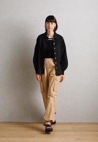 Une personne porte une chemise noire à rayures oversize sur un top noir, associée à un pantalon cargo beige avec des poches et des sandales noires.