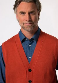 Oranžová pletená vesta s hlubokým V-výstřihem, zdobená černými knoflíky, přes modrou denimovou košili. Hladká textury a přiléhavý střih.