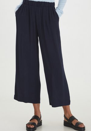 Mujer con pantalones anchos negros de pierna ancha y cintura elástica, y sandalias negras con plataforma, con las manos en los bolsillos.