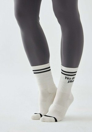 Nogi w ciemnoszarych legginsach, noszące białe skarpety za kostkę z czarnymi paskami i napisem "No drama please", stojące na czubkach palców na białej powierzchni.