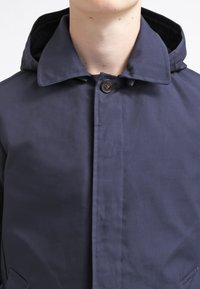 Giacca a vento blu navy con chiusura a bottoni, caratterizzata da una texture liscia e un design classico. Include un colletto ripiegato e tasche laterali.