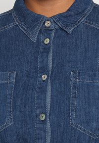 Dark blue denim shirt with a pointed collar, vertical row of buttons, and two chest pockets with visible stitching.