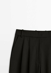 Pantalons plissés noirs avec une texture lisse, taille plate et poches latérales. Conçus pour une silhouette structurée.
