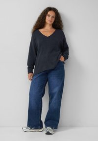 Donkergrijze v-hals trui met verlaagde schouders, gecombineerd met losse blauwe spijkerbroeken. De outfit wordt aangevuld met zilveren sport sneakers.