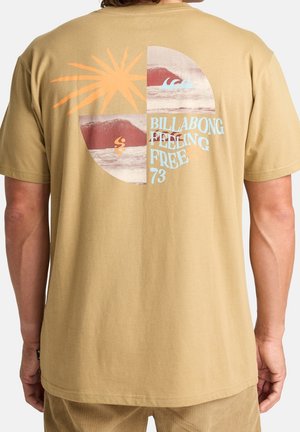 Beige Hemd mit einem segmentierten Kreisgrafik, der Sonne, Wellen, Berge und den Text "BILLABONG FEELING FREE 73" auf der Rückseite zeigt.