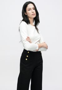 Top alb cu mâneci lungi combinat cu pantaloni negri cu talie înaltă, accesorizați cu nasturi aurii și picioare largi, pe un fundal deschis.