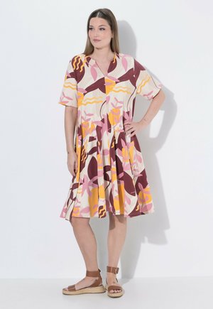 Mujer con vestido de manga corta y largo hasta media pierna con un patrón floral abstracto en rosa, amarillo y granate, y sandalias marrones con correa en el tobillo.