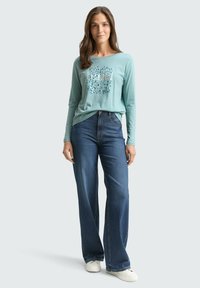 Woman wearing a light blue long-sleeve shirt with “beautiful” text, dark blue wide-leg jeans, and white sneakers, standing against light background.