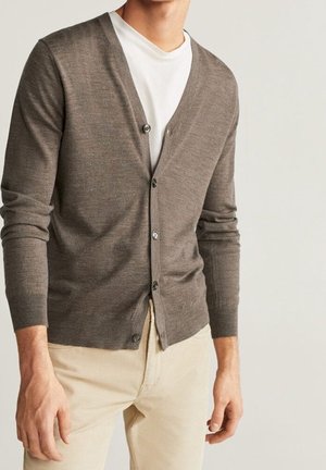 Homme portant un cardigan marron boutonné sur un t-shirt blanc avec un pantalon beige, debout devant un fond clair uni.