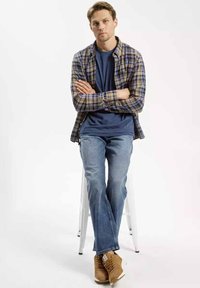 Koszula w kratę w odcieniach flaneli na granatowej koszulce, w połączeniu z jasnoniebieskimi dżinsami i brązowymi trampkami. Model siedzi na białym stołku.