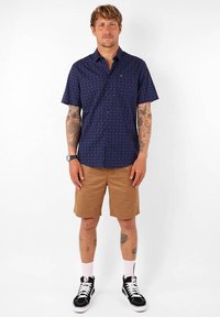 Chemise à manches courtes bleu marine à boutons avec un motif géométrique blanc, assortie à un short beige. Des baskets montantes noires et des chaussettes blanches à la cheville complètent le look.