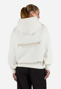 Biely oversized mikina z mäkkého materiálu, s kapucňou a rebrovanými manžetami. Na zadnej strane je logo "PEGADOR" v béžovej farbe.