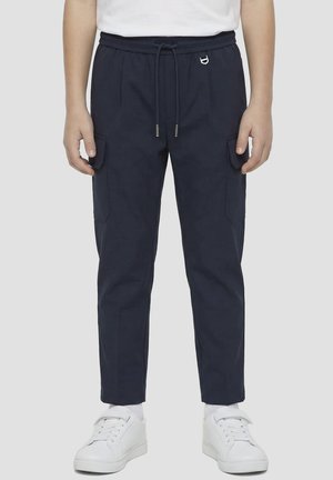 Dijete u tamnoplavim cargo hlačama s vezicom u struku i bijelim tenisicama na svijetlosivoj pozadini.