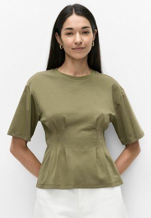 Vrouw met lang donker haar die gouden creolen draagt, een olijfgroene nauwsluitende top met korte mouwen en een witte broek, staand met handen achter de rug.