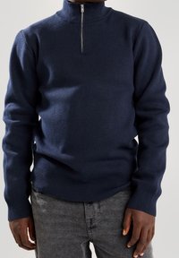 Pull en maille bleu marine avec col montant et fermeture éclair à l'avant, ourlet et poignets côtelés. Présente une surface texturée et une silhouette ajustée.
