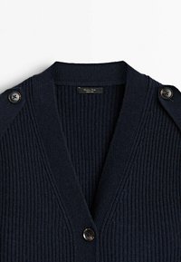 Cardigan en maille côtelée bleu marine foncé avec col en V, devant boutonné et épaulettes boutonnées, étiquette indiquant Fabriqué en Chine.