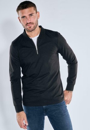 Schwarzes Poloshirt mit langen Ärmeln und Reißverschlusskragen aus strukturiertem Material. Kombiniert mit blauen Jeans. Merkmale ein dezentes Logodetail am Saum.
