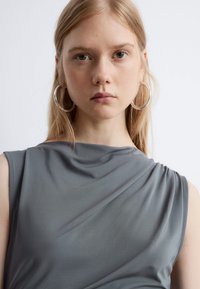 Graues, ärmelloses Top mit drapiertem Design, das aus weichem, glattem Stoff mit subtiler Textur besteht. Große silberne Creolen betonen den Look.