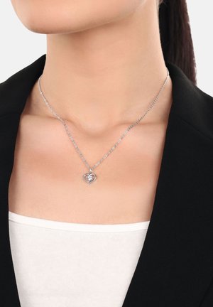 Colgante en forma de corazón de plata con una piedra preciosa clara en el centro, llevado alrededor del cuello con un blazer negro y una blusa blanca.
