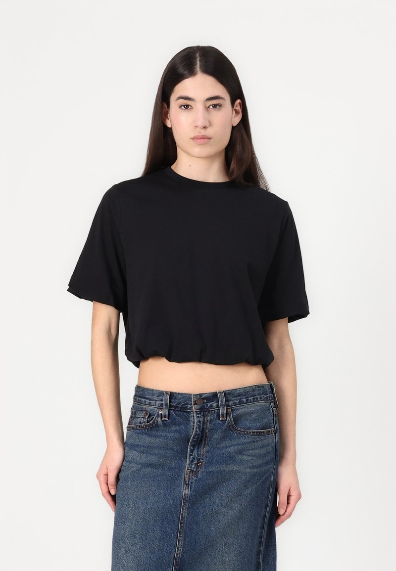 Fekete, pamutból készült, rövid ujjú cropped póló. A modell egy magas derekú farmer szoknyával párosítja, amely gombos záródással rendelkezik.