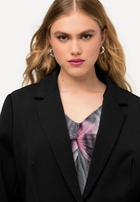 Blazer negro con solapas de muesca sobre una blusa con un patrón abstracto en rosa y gris. La modelo tiene el cabello rubio ondulado y pendientes de aro plateados.