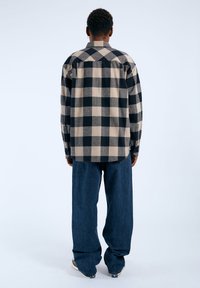 Camisa de cuadros con cuadrados negros y beige, con diseño de botón en la parte delantera y mangas largas, combinada con pantalones de mezclilla azul sueltos.