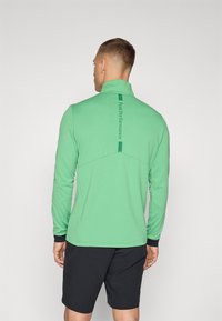 Grüner Pullover mit langen Ärmeln, hohem Kragen und Detail an der Rückennaht. Verfügt über schwarze Akzente an den Bündchen und ein Logo entlang der Wirbelsäule.