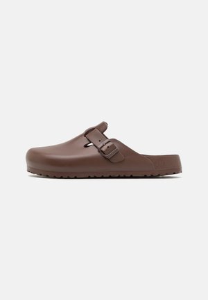 Zoccolo marrone slip-on realizzato in plastica liscia con suola testurizzata, dotato di una cintura regolabile e punta arrotondata.