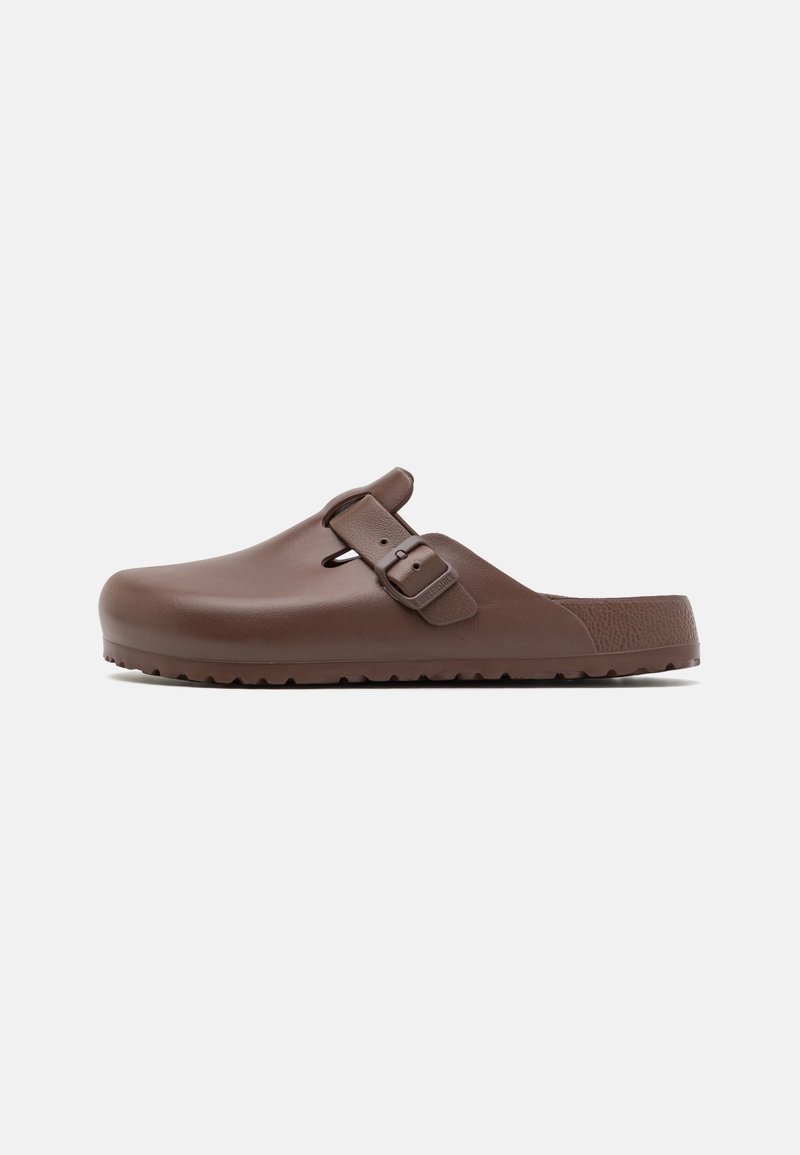 Zoccolo marrone slip-on realizzato in plastica liscia con suola testurizzata, dotato di una cintura regolabile e punta arrotondata.