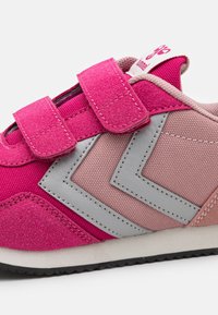 Baskets de sport rose et grise avec une tige en daim texturé, des sangles Velcro et un design en zigzag sur le côté. Semelle en caoutchouc.