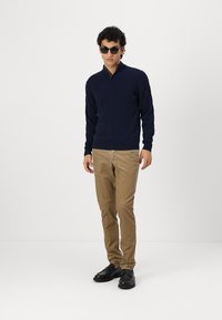 Pull en maille bleu marine avec un col montant, associé à un pantalon marron clair et des chaussures noires. Le modèle porte également des lunettes de soleil noires.
