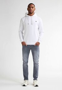 Bílá mikina s kapucí a stahovacími šňůrkami, logo na hrudi, kombinovaná se světle modrými džíny a bílými teniskami. Jednoduchý design, bez viditelných vzorů nebo ozdob.