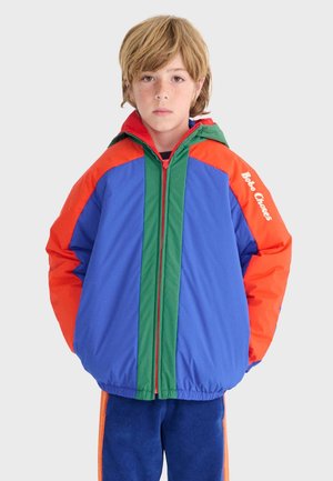 Enfant aux cheveux blonds portant une veste à capuche bleue, orange et verte, mains dans les poches, debout devant un fond uni.